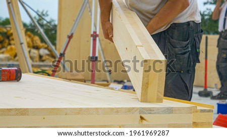 CLOSE UP, DOF: Unrecognizable male builder picks up a CLT beam from a workbench at an unfinished housing project. Contractor carries a glued laminated board across the bustling construction site Royalty-Free Stock Photo #1961969287