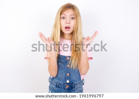 Surprised terrified beautiful caucasian little girl wearing jeans overall over white background Gestures with uncertainty, stares at camera, puzzled as doesn't know answer on tricky question.
