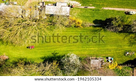 A view of a large ride along lawn mower cutting the grass of a large garden, at the back of a small property. The sun shining above during spring time.