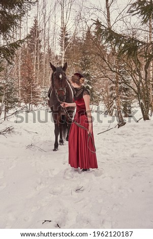 A girl with blonde hair in a vintage red dress stands next to a horse in a winter forest.