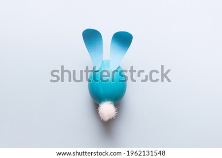 Easter composition with one cartoon stylized blue egg with paper ears and white fluffy tail on blue background.