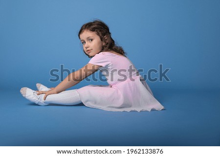 Side portrait of a little ballerina performing stretching legs, sitting over blue background with space for text
