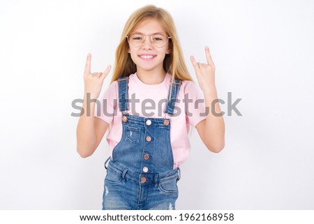 beautiful caucasian little girl wearing jeans overall over white background making rock hand gesture and showing tongue
