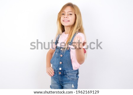 beautiful caucasian little girl wearing jeans overall over white background inviting to come with hand. Happy that you came