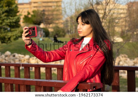 Green-eyed Latina teen taking a selfie with her mobile in the park with a red check