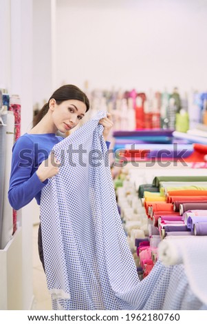 Woman customer or buyer in fabrics store matching textile cloth on herself, applying it to the body in front of the mirror. Tailoring, seamstress or atelier concept. High quality vertical image