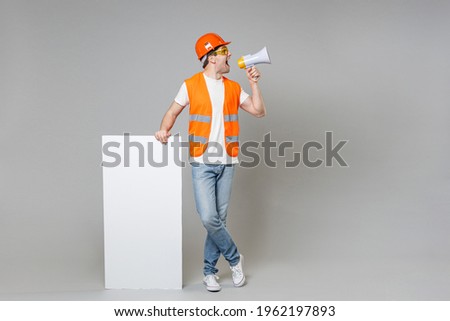 Full length employee man in orange vest hardhat hold blank sign board, place for text scream aside in megaphone isolated on grey background studio. Instruments for renovation apartment. Repair concept