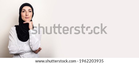Young woman dressed in a white shirt and hijab holds a hand to the chin on a light background. Banner.