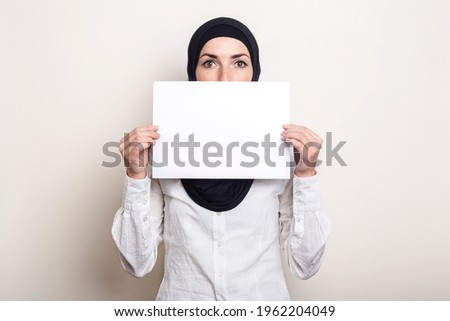 Young Muslim woman in a white shirt and hijab covers her face with a blank sheet of paper. Place for your text. Banner.