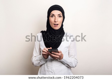 Muslim young woman with a surprised face in a hijab holds a phone in her hands on a light background. Banner. Looks at the camera.