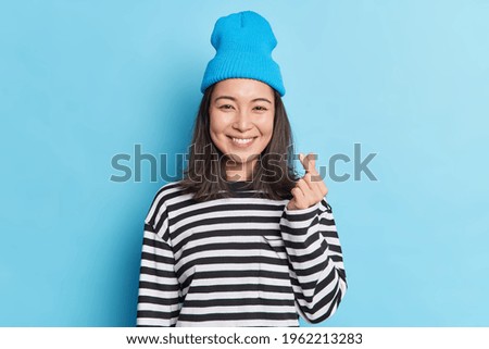 Waist up shot of pretty Asian woman with dark hair makes korean heart with fingers wears hat striped jumper smiles pleasantly isolated over blue background expresses love. Body language concept Royalty-Free Stock Photo #1962213283