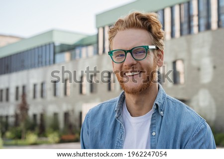 Young successful student looking at camera standing in university campus, education concept. Portrait of handsome smiling Irish man wearing stylish eyeglasses posing for pictures outdoors 	