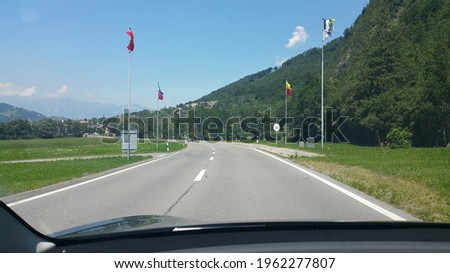 A shot on the move from the windshield of an electric car with Switzerland-Liechtenstein border in front of it in a sunny summer day. POV first person view shot on a mountain road.