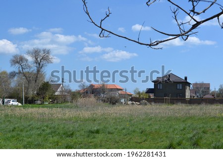 houses in the village in front of them is a large beautiful field with green grass trees grow next to the houses they bloom above the houses a bright blue sky with clouds spring