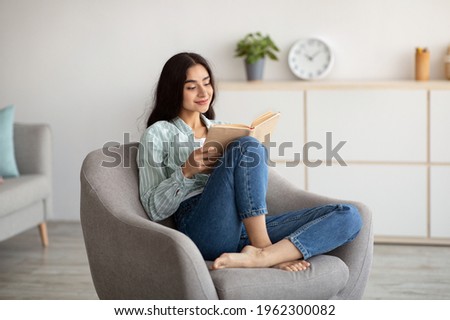 Stay home pastimes. Millennial Indian woman sitting in cozy armchair with open book indoors. Beautiful young lady reading exciting story, enjoying lazy morning, having relaxing weekend Royalty-Free Stock Photo #1962300082