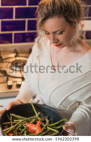 Young blond woman cooks in the kitchen. Delicacy. Seafood. Shrimp, asparagus in a pan.