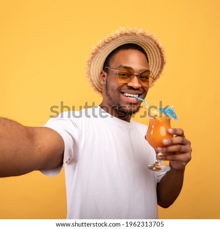 Handsome black guy in summer outfit drinking exotic cocktail and taking selfie on yellow studio background. Happy young man making smartphone photo of himself during tropical vacation