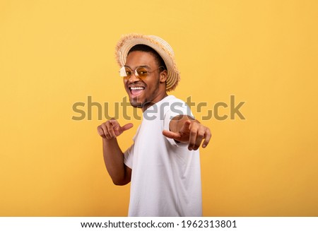 Happy black guy in casual summer outfit, straw hat and sunglasses pointing at you on yellow studio background. Positive African American man enjoying summertime, having fun