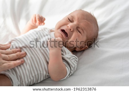 Closeup Portrait Of Crying Little Newborn Baby In Bodysuit Lying On Bed At Home, Upset Infant Child Suffering Colics Or Gas Problems, Loving Mother Making Tummy Massage To Small Kid With Two Hands Royalty-Free Stock Photo #1962313876
