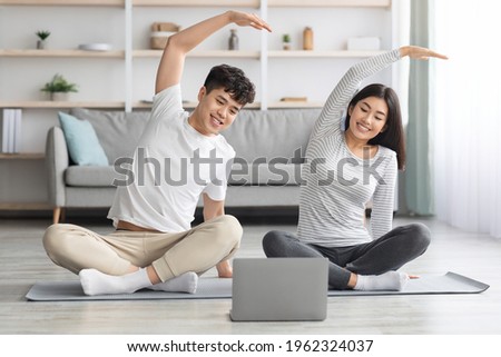Cheerful asian family practicing yoga at home, stretching on fitness mat, looking at laptop screen, having online class, living room interior, copy space. Sporty black couple exercising together Royalty-Free Stock Photo #1962324037