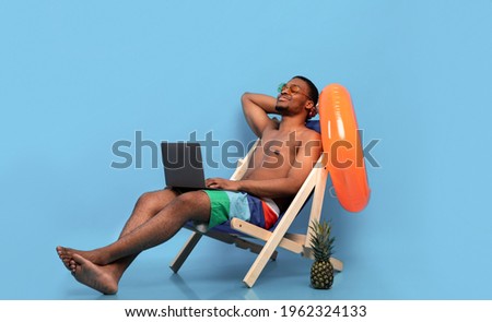 Relaxed black guy chilling in lounge chair with laptop computer, working online from tropical paradise, blue background. Cool young man on summer vacation watching movie or talking to friend on pc
