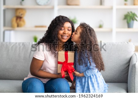 Mother's Day celebration. Lovely little black girl giving mom wrapped holiday gift and kissing her on cheek at home. Cute daughter congratulating her mommy with birthday. Family traditions concept