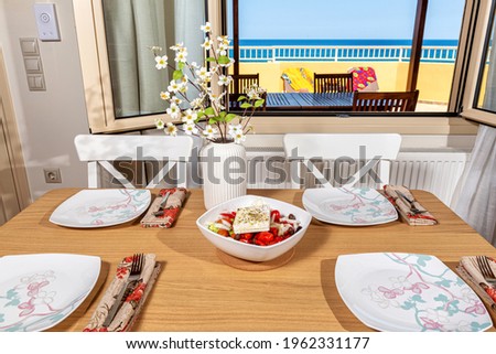 Wooden table served for lunch, a plate of fresh greek salad on it. At the blurred background open window with the virew to the sunbathing terrace and seascape. 