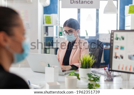 Woman entrepreneur working on laptop in new normal office with coworkers keeping social distnacing behind plastic shiled, wearing face mask as safety prevention during cornavirus flue outbreak. Royalty-Free Stock Photo #1962351739