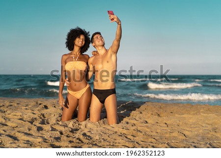 Summertime romantic selfie on the beach. Interracial couple having fun using smartphone and taking pictures kneeling on the sand.