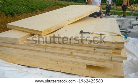 CLOSE UP: Cross-laminated timber wall panels lie on the ground next to a group of unrecognizable workers. A stack of CLT wall panels lie uncovered at a construction site of a modern housing project. Royalty-Free Stock Photo #1962425560