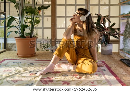 Satisfied woman wearing headset listen to music relaxed after remote work day at home. Cute female gardener sit on floor with closed eyes dreaming. Young casual girl small business owner relaxation Royalty-Free Stock Photo #1962452635