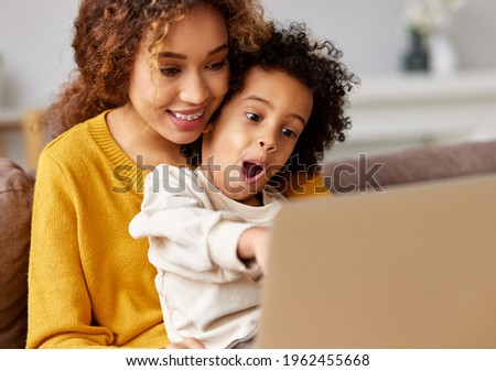 Excited little boy son watching video or cartoons on laptop with young smiling mother, pointing with finger at computer screen and keeping mouth opened, a mom and kid spending leisure time at home