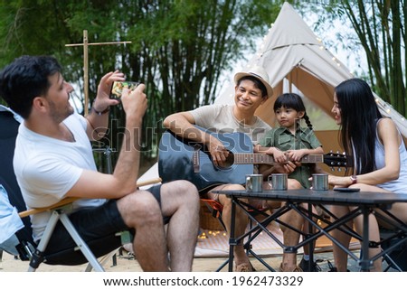 Caucasian father using cellphone taking a picture of grandfather holding guitar with little daughter and Asian mother standing and sitting next to him while enjoyed camping in the forest after hiking.