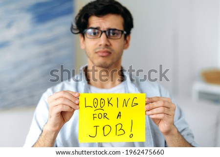 Blurred sad unemployed young clever hispanic man with glasses, student, stands with sign with the inscription looking for a job, hopes to get a dream job, asks for support, looks at camera