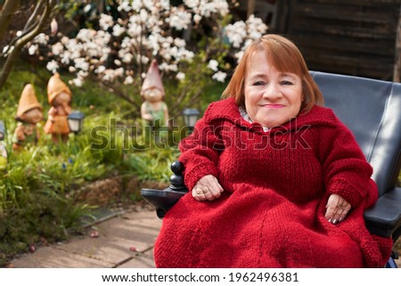 a small woman with brittle bone disease is sitting in a wheelchair on the terrace in her garden - she is wearing a pretty red self knitted dress and looks very happy and friendly Royalty-Free Stock Photo #1962496381