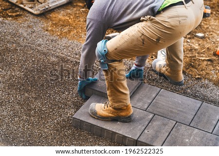 Landscaping paver worker laying paving stones on sandy ground of construction patio yard site in spring summer. Contractor wearing safety protective cloth, gloves and knee pads for installation work.  Royalty-Free Stock Photo #1962522325