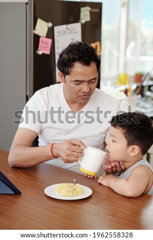 Father giving his little son a cup of water when they are eating breakfast and watching animated film on tablet computer