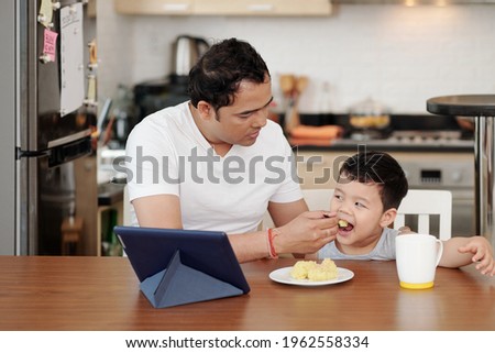 Father feeding his little son with mashed potato when they are watching cartoon on tablet computer
