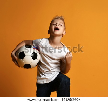 Happy schoolboy, teenager, soccer fan in white blank t-shirt and jeans stands holding soccer ball in hand and celebrates Goal over yellow background