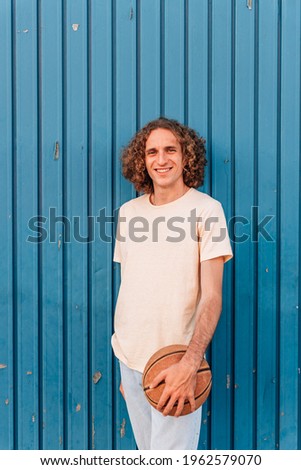 vertical portrait of a young caucasian man looking at camera smiling and holding a basketball. He has long red hair and wears casual summer clothes. It is leaning on a blue metal door