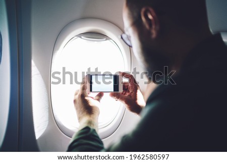 Side view of unrecognizable bearded man with eyeglasses travelling on plane and taking picture of view outside through window on sunny day