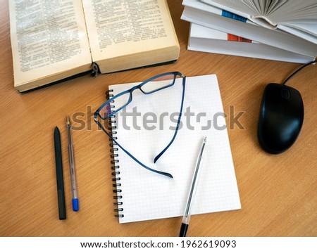Glasses and a pen are on a blank sheet of notebook. There are open and closed books, a computer mouse and pens nearby. The concept of education, e-learning.