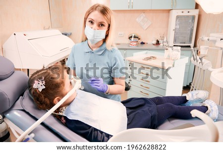 Young woman stomatologist in medical face mask looking at camera and holding dental instruments while little girl lying in dental chair with inhalation sedation. Concept of sedation dentistry. Royalty-Free Stock Photo #1962628822