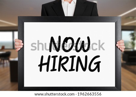 Businessman holds a big signboard with now hiring message. Business recruitment or employment concept.

