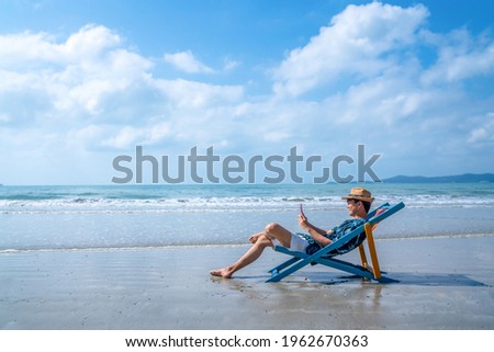 Asian man lying on beach chair using smartphone chatting or online shopping during travel at the beach on summer holiday vacation. People enjoy outdoor lifestyle with gadget device and online network. Royalty-Free Stock Photo #1962670363
