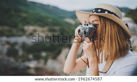 young blonde girl in summer hat takes photo on retro camera on background panorama horizin mountain landscape, hipster tourist enjoys hobby of photographer  leisure in summer nature empty space