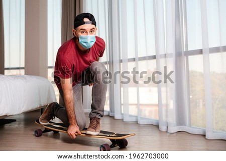 A young man in a medical mask poses while standing on a longboard. Windows in the background. Indoors. Concept of quarantine and activity at home.
