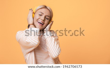 Charming woman is listening to music on headphones posing on a yellow studio wall advertising something