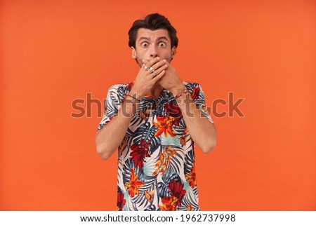 Scared shocked young man with bristle in colourful shirt covered his mouth by hands and feels frightened over orange background