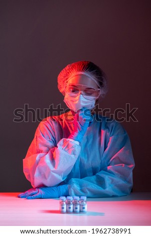 Vaccine risk. Covid-19 inoculation. Clinical trial. Lab research. Pensive doubtful female scientist in ppe face mask gloves looking at dose vials in red blue neon light isolated on dark.